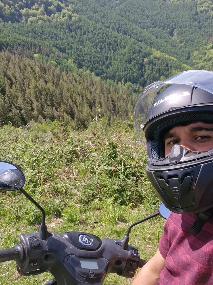 Selfie of Abhiram in a motorcycle helmet on a scooter, above a valley of dense green forest