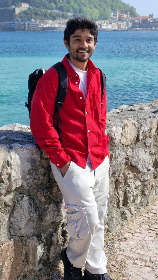 Abhiram leaning on a stone sea wall by turquoise water, with a hillside town behind him