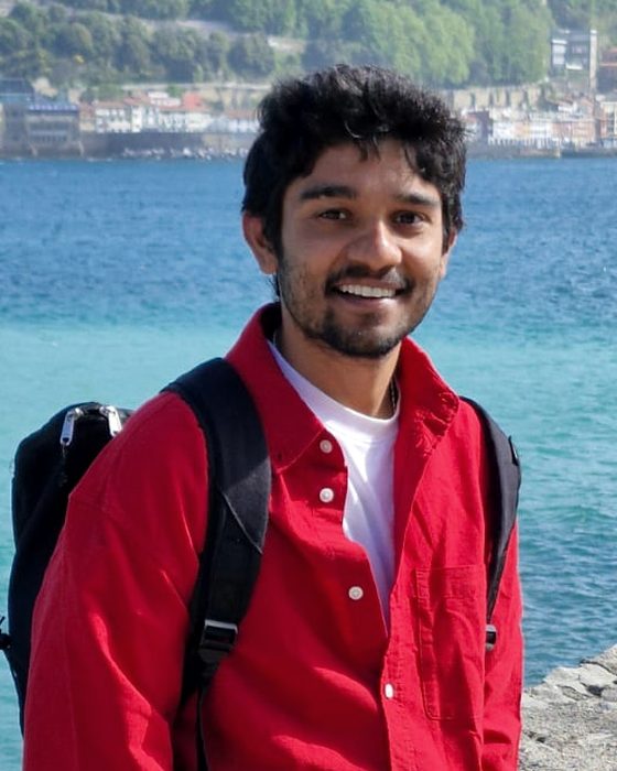 Portrait of Abhiram smiling, in a red shirt, with the sea behind him