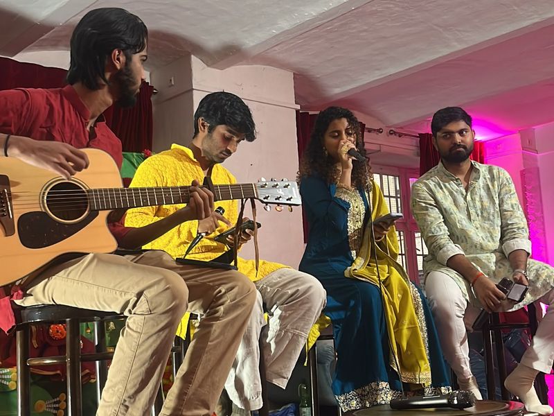 Abhiram in a yellow kurta on stage with three other musicians at an acoustic music evening, a guitarist beside him and a singer at the microphone