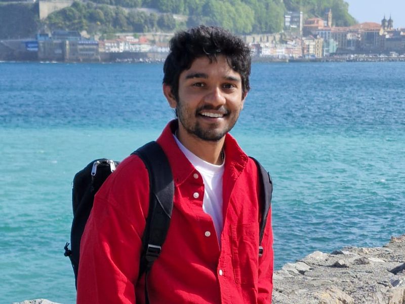 Abhiram smiling by turquoise sea, with an old harbour town on the hillside behind him